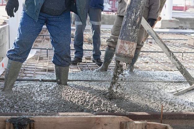 Trabajadores de la construcción vierten piso de concreto en taller industrial Piernas con botas en concreto Presentación de concreto para verter el piso Obras monolíticas de concreto