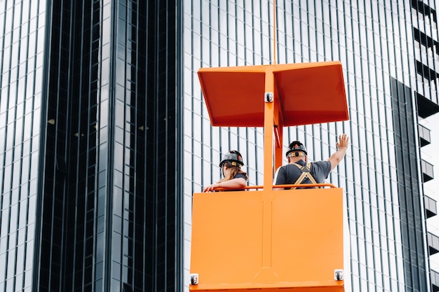 Los trabajadores en una cuna de construcción se suben a una grúa a un gran edificio de vidrio. La grúa levanta a los trabajadores en el asiento del automóvil.