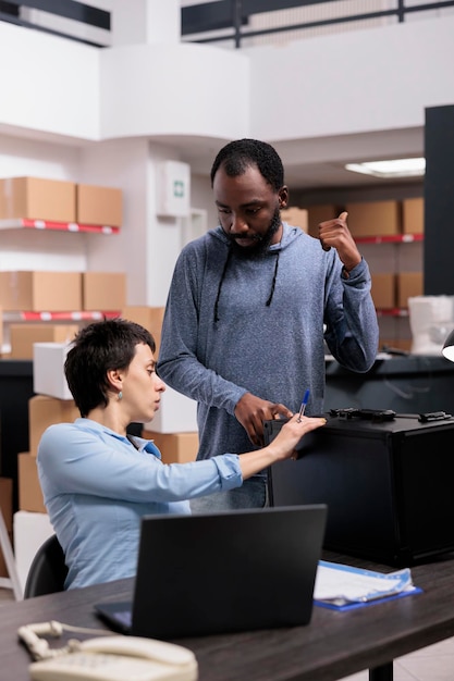 Los trabajadores de entrega del almacén revisan la caja metálica mirando el pedido del paquete mientras trabajan en la producción de empaques en el almacén. Equipo multiétnico que analiza la logística de envío en una computadora portátil