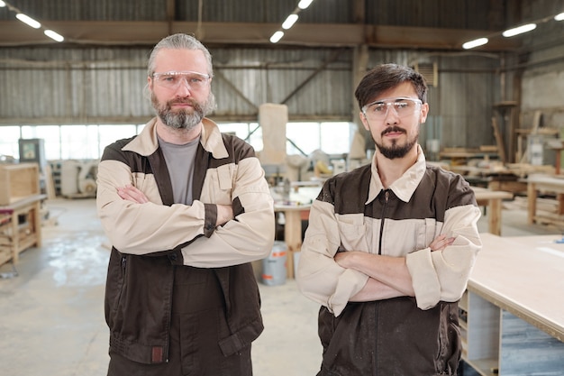 Trabajadores masculinos en ropa de trabajo y anteojos protectores usando una máquina de pulir para procesar la superficie de la tabla de madera por un banco de trabajo