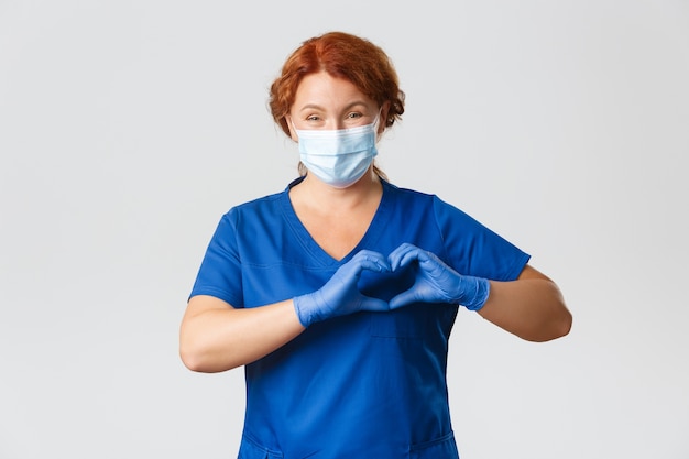 Trabajadores médicos, pandemia de covid-19, concepto de coronavirus. Hermosa doctora sonriente pidiendo a las personas que se queden en casa seguras, que usen mascarilla y guantes, que muestren el gesto del corazón, que atiendan a los pacientes en la clínica.