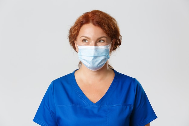 Trabajadores médicos, pandemia de covid-19, concepto de coronavirus. Pensativo sonriente doctora, enfermera de mediana edad o médico con mascarilla y matorrales mirando la esquina superior izquierda, fondo gris.