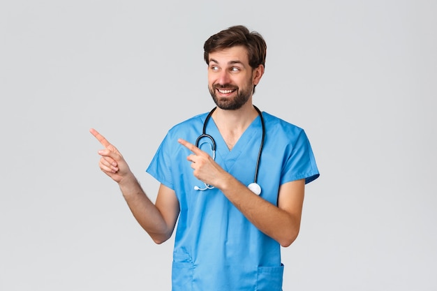 Trabajadores de la salud, concepto de brote de pandemia y coronavirus. Médico barbudo entusiasta en matorrales, estetoscopio, señalando con el dedo hacia la izquierda y sonriendo, leyendo promo o publicidad, fondo gris