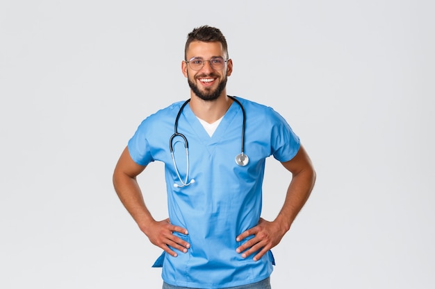 Trabajadores de la salud, medicina, covid-19 y concepto de autocuarentena pandémica. Entusiasta médico guapo, enfermero con gafas y batas azules, sonriendo disfrutando de trabajar con pacientes de la clínica