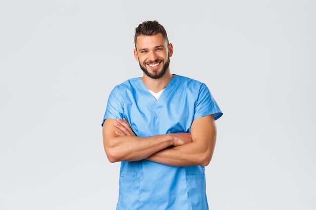 Trabajadores de la salud, medicina, covid-19 y concepto de autocuarentena pandémica. Sonriente médico guapo, hispano, enfermero en matorrales azules, pecho de brazos cruzados, cámara de risa alegre.