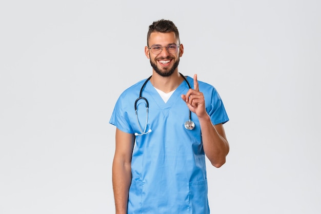 Trabajadores de la salud, medicina, covid-19, concepto de cuarentena pandémica. Amistoso trabajador del hospital sonriente, enfermera o médico con batas azules, gafas, apuntando con el dedo hacia arriba, brindan información a los pacientes.