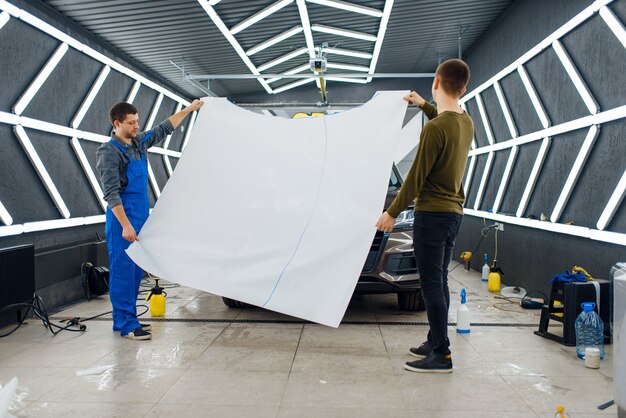 Los trabajadores varones tienen plantilla de capó de película de protección de coche. Instalación de revestimiento que protege la pintura de automóvil de arañazos. Vehículo nuevo en garaje, procedimiento de puesta a punto.