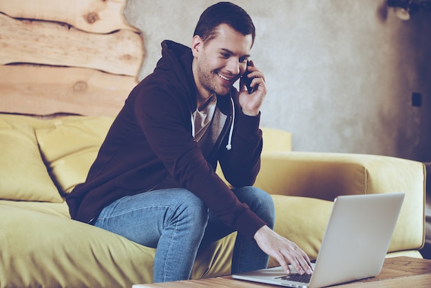 Trabajando desde casa con mucho gusto. Joven alegre usando su computadora portátil y hablando por teléfono móvil con una sonrisa mientras está sentado en el sofá en casa