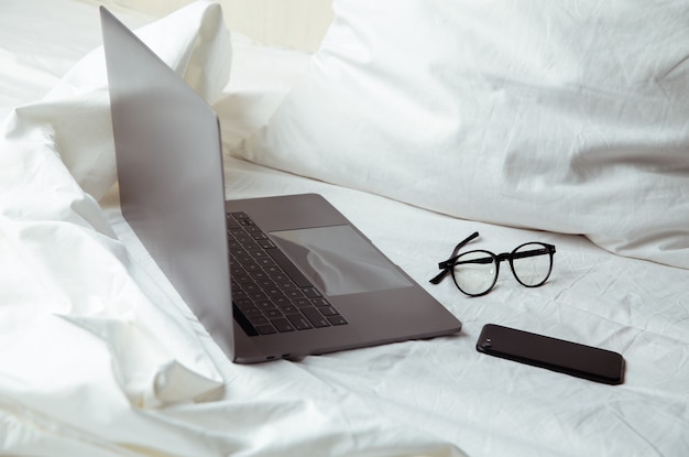 Trabajando desde casa. Portátil y gafas en la cama blanca