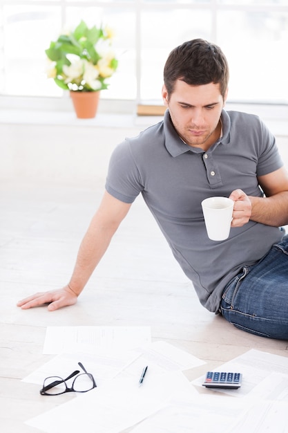 Trabajando en casa. Vista superior del joven confiado sosteniendo una taza y mirando los documentos mientras está sentado en el suelo de su apartamento