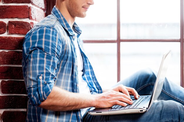 Trabajando en una computadora portátil. Vista lateral del joven que trabaja en la computadora portátil mientras está sentado en el alféizar de la ventana