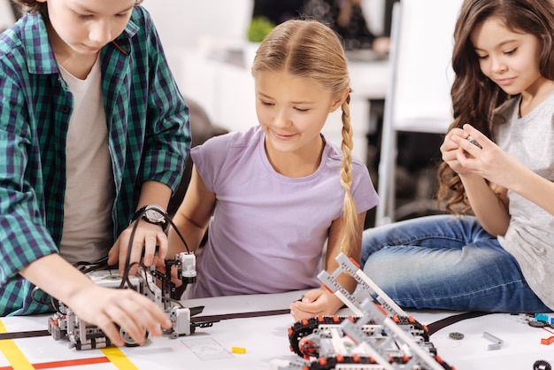 Trabajando con detalles. Niños atentos encantados trabajadores sentados en el laboratorio de robótica y reparando dispositivos cibernéticos mientras tienen una lección de ciencias