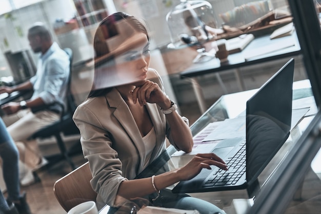 Trabajando duro. Vista superior de la mujer joven moderna que usa la computadora mientras trabaja detrás de la pared de vidrio en la oficina