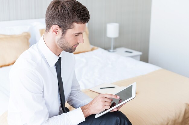 Trabajando en la habitación del hotel. Hombre de negocios joven confiado en camisa y corbata trabajando en tableta digital mientras está sentado en la cama en la habitación del hotel