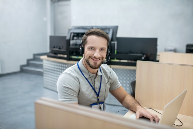 Trabajando con información. Especialista masculino sonriente en auriculares con micrófono detrás del portátil en una habitación especialmente equipada