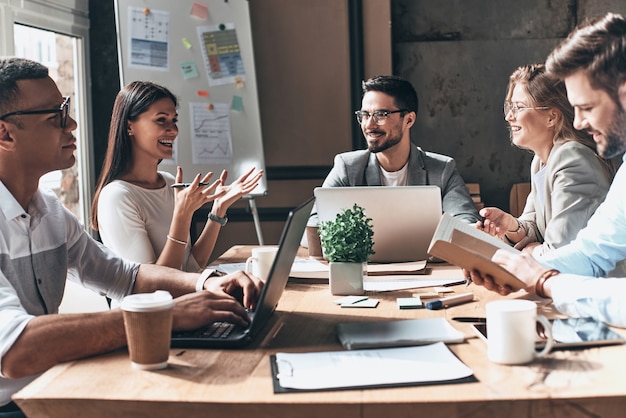 Trabajando juntos. Grupo de jóvenes modernos en ropa casual elegante discutiendo negocios y sonriendo mientras está sentado en la oficina creativa