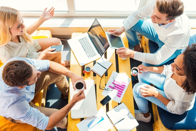 Trabajando juntos para obtener mejores resultados. Vista superior de cuatro jóvenes felices trabajando juntos mientras están sentados en el escritorio de madera en la oficina