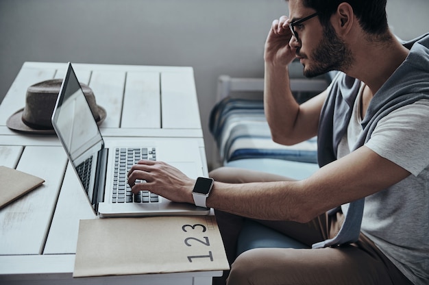 trabajando para lograr el éxito. Hombre moderno joven pensativo ajustando sus gafas y usando la computadora