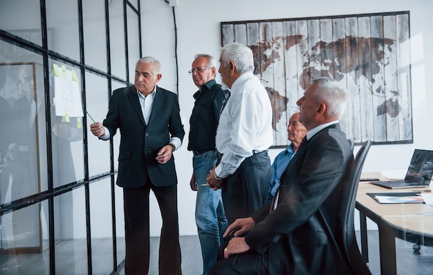 Trabajando con pegatinas sobre vidrio. Equipo envejecido de arquitectos de negocios de edad avanzada tiene una reunión en la oficina.