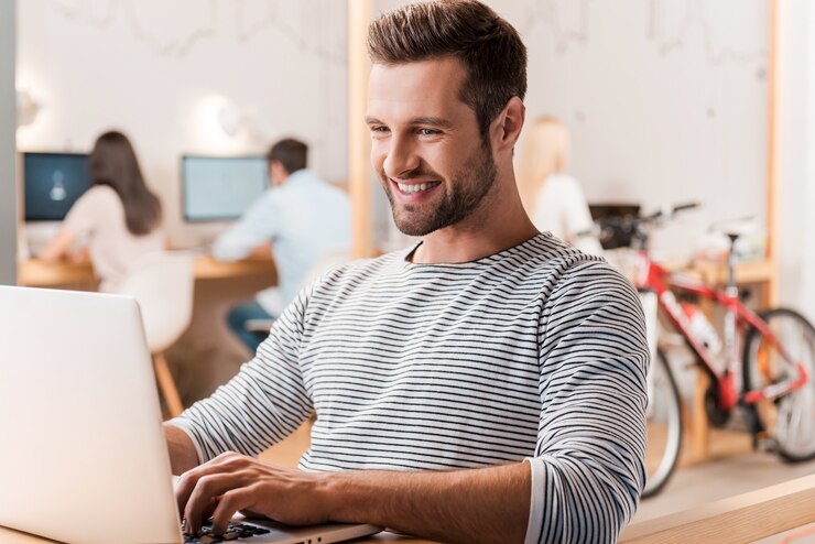 Foto trabajando con placer. apuesto joven trabajando en la computadora portátil y sonriendo mientras sus colegas trabajan en segundo plano