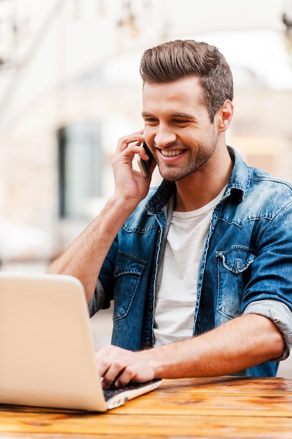 Trabajando con placer. Hombre joven feliz que trabaja en la computadora portátil y hablando por el teléfono móvil mientras está sentado en la mesa de madera al aire libre