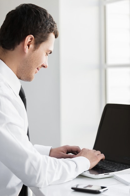 Trabajando con placer. Vista lateral del joven sonriente en ropa formal que trabaja en la computadora portátil mientras está sentado en su lugar de trabajo