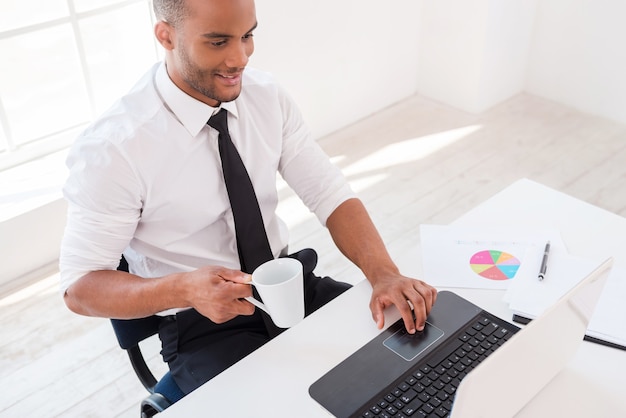 Trabajando con placer. Vista superior del hombre africano joven confiado en camisa y trabajando en la computadora portátil