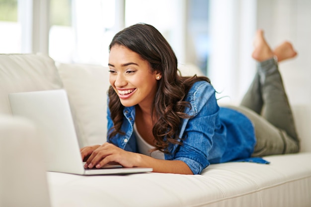 Trabajando en su blog Captura recortada de una joven atractiva usando su computadora portátil mientras está acostada en el sofá