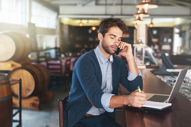 Trabajando en su blog en su bar favorito Captura recortada de un apuesto joven usando su computadora portátil y su teléfono celular en un bar