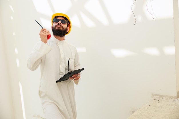 Trabajando en tableta y walkie talkie Hombre árabe Ingeniero civil de construcción usa una tableta digital y usa casco de seguridad trabajando en el sitio de construcción