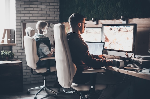 Trabajando con tecnologías modernas. Vista trasera del equipo de jóvenes empresarios trabajando y mirando el monitor mientras está sentado en el escritorio en la oficina creativa