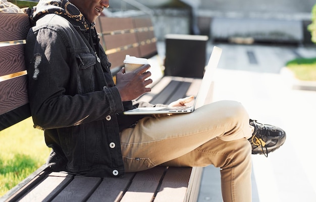 Trabajando usando una computadora portátil Vista de cerca del hombre afroamericano con chaqueta negra al aire libre en la ciudad sentado en un banco