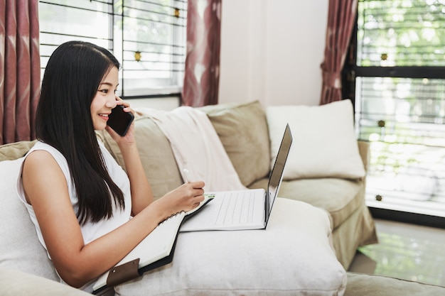 Trabajar desde casa y el concepto de compras en línea, mujer asiática de negocios feliz con teléfono móvil y computadora portátil con computadora portátil en el sofá en la sala de estar en casa