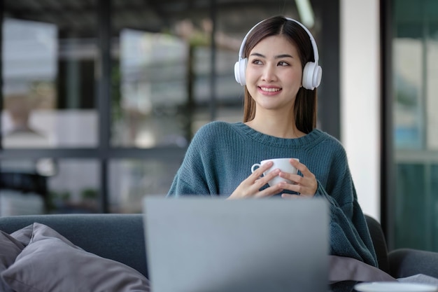 Trabajar en casa joven mujer asiática trabajando en la computadora portátil mientras está sentada y sostiene la taza de café en casa concepto de estilo de vida de la mañana
