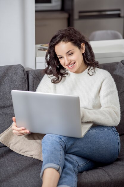 Trabajar desde casa. Una mujer joven con un suéter blanco sentado en el sofá con una computadora portátil