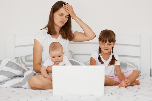 Foto trabajar desde casa con niños pequeños, madre adulta joven tratando de trabajar en la computadora portátil mientras cuida niños, madre estresante tratando de realizar múltiples tareas mientras está sentada en la cama con sus hijos.