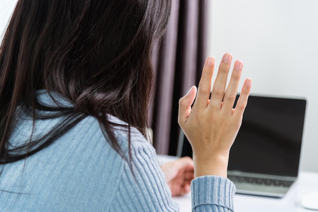 Trabajar desde casa, la parte posterior de una joven empresaria asiática llamada de videoconferencia o una reunión en tiempo real en línea levantan la mano para saludar en la computadora portátil frontal pone en cuarentena la enfermedad coronavirus, Enfoque en la computadora portátil