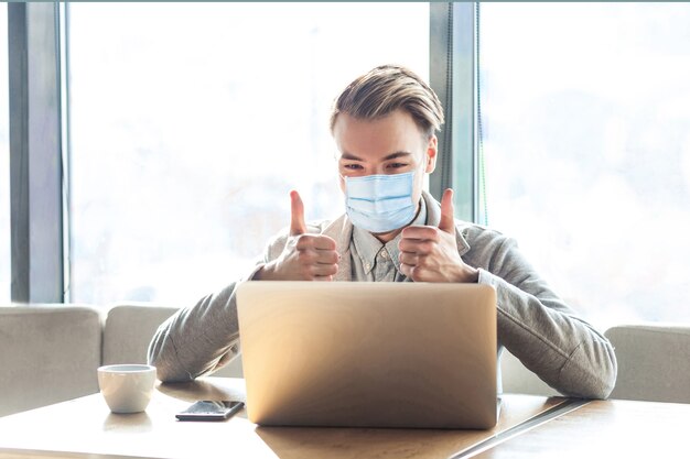 Trabajar en casa seguro en tiempo de cuarentena. Joven satisfecho con máscara médica quirúrgica, sentado, mirando la pantalla del portátil con los pulgares hacia arriba. interior, estilo de vida autónomo, trabajo a distancia, asistencia sanitaria.