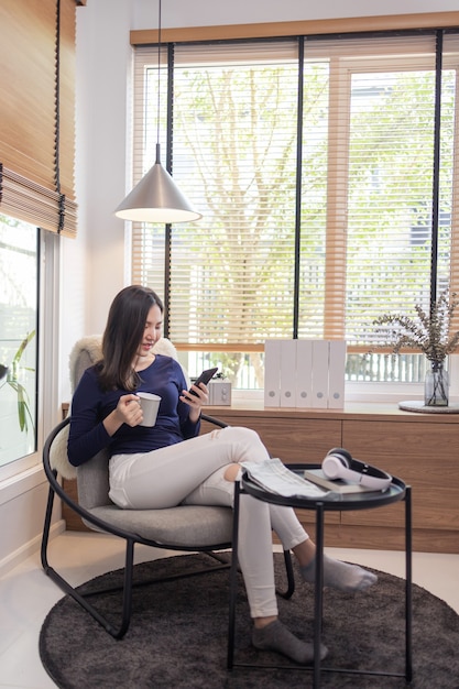 Trabajar desde el concepto de casa una mujer segura de sí misma sentada en una silla moderna sostiene una taza de café y otra mano escribiendo un teclado en su teléfono inteligente.