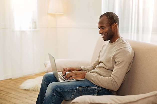 Trabajar por cuenta propia. Feliz sonriente hombre afroamericano escribiendo en la computadora y mirando la pantalla mientras está sentado en el sofá