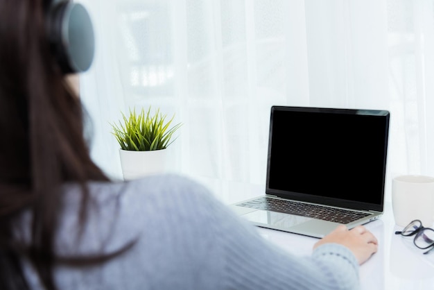 Trabajo desde casa, llamada de videoconferencia en la parte posterior de una joven empresaria asiática o reunión presencial en línea en la computadora portátil frontal pone en cuarentena el coronavirus de la enfermedad o COVID-19 en la oficina del hogar, enfoque en la computadora portátil