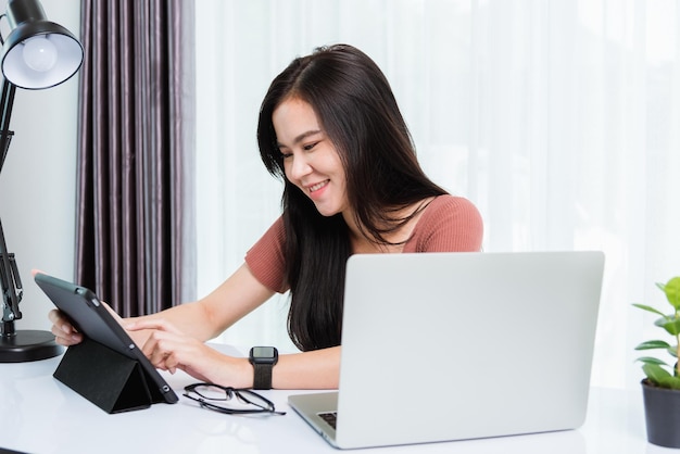 Trabajo desde casa, Sonriente feliz mujer de negocios asiática, joven y hermosa mujer sentada en el espacio de trabajo del escritorio, use una tableta inteligente moderna y tecnología de computadora portátil, videollamadas, conferencias para el equipo en la oficina en casa