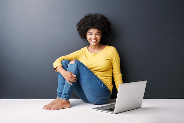Trabajo de clase en línea y retrato de mujer negra con maqueta de portátil y felicidad en el suelo en el fondo del estudio Investigación informática o estudiante universitario escribiendo en el teclado para un proyecto de trabajo o aprendizaje electrónico