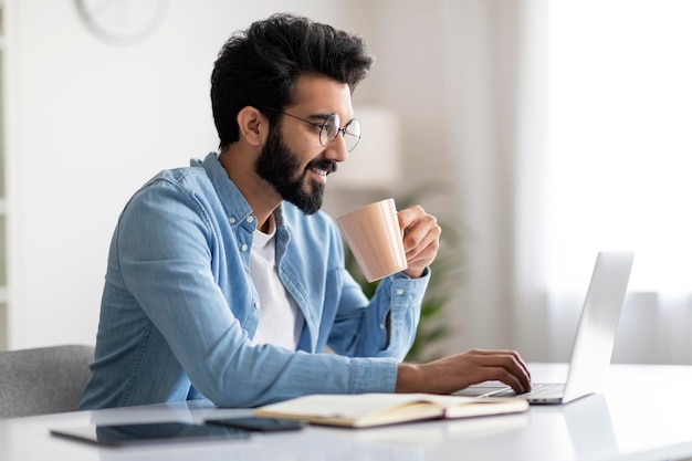 Trabajo a distancia apuesto trabajador independiente indio bebiendo café y trabajando con una computadora portátil