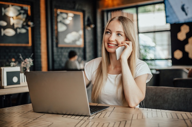 Trabajo a distancia Joven mujer rubia caucásica sentada en un café con una computadora portátil y hablando por teléfono