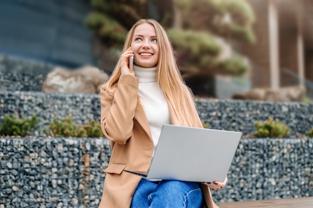 Trabajo a distancia. Mujer de negocios joven rubia sentada en un banco con un teléfono móvil y una computadora portátil, hablando y trabajando