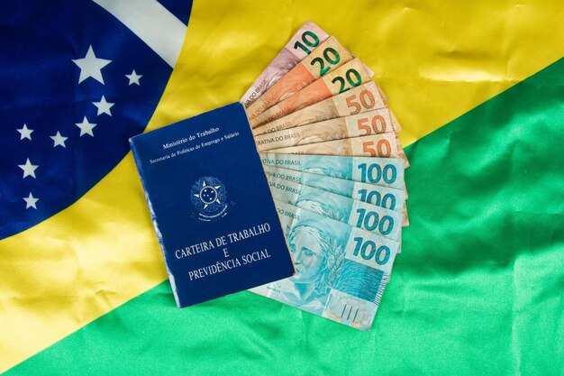 Trabajo de documentos brasileños y seguridad social Carteira de Trabalho e Previdencia Social con billetes de dinero brasileños y bandera brasileña en el fondo