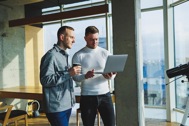 Trabajo de dos colegas masculinos de pie juntos en la oficina mientras intercambian ideas con una computadora portátil en la sala de reuniones y analizan esquemas de trabajo Trabajar en una oficina moderna y espaciosa