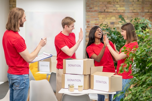 Trabajo en equipo. Jóvenes voluntarios alegres en camisetas rojas que trabajan en las instalaciones del centro de caridad empacando cajas de donación