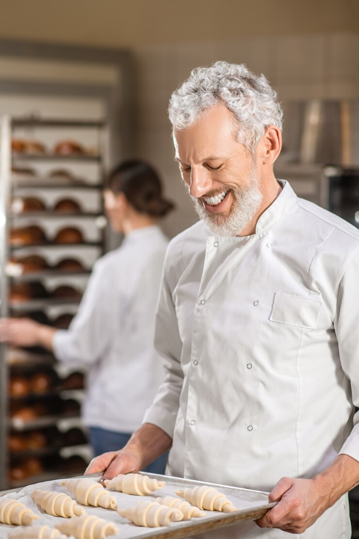 Foto trabajo favorito. hombre barbudo gris feliz mirando la bandeja de croissants en las manos y la mujer ocupada detrás en la panadería